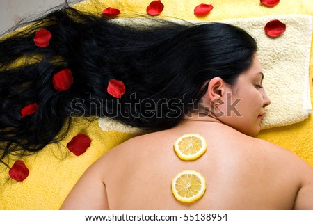 Top view of beautiful woman with long hair lying on massage table and having  slice lemon treatment on her back and rose petals in hair