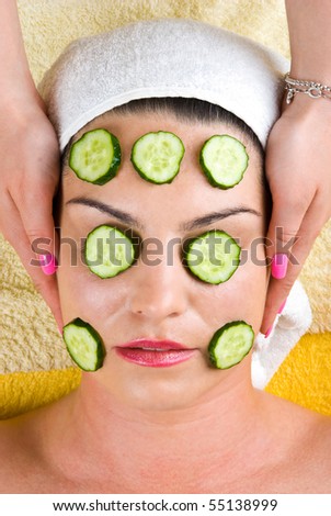 Beautician making a facial mask with slice  cucumber to a young woman and holding her hands on client head