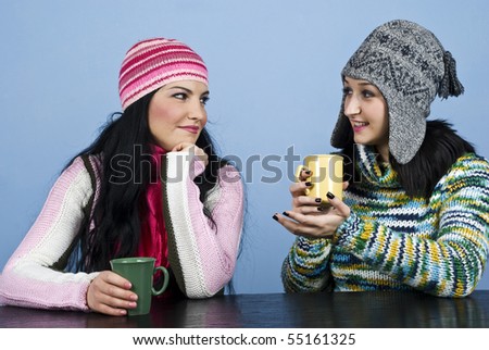 Two young women in winter clothes sitting at table and having a funny discussion while enjoying a cup of hot drink together