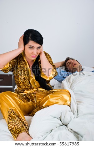 Couple in bed with woman awake and nervous because can\'t sleep and holding hands on ears while man sleeping and snoring