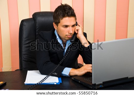 Busy business man in office sitting on chair at desktop talking by telephone and typing on laptop