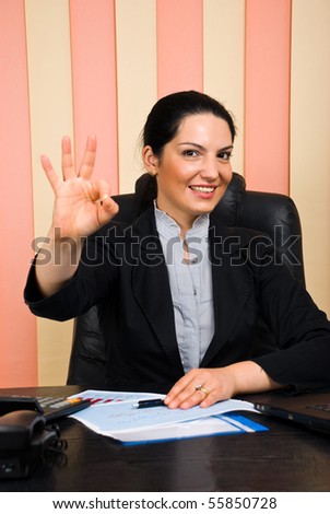 Beautiful happy business woman formal wear sitting at desk in office and showing okay hand sign