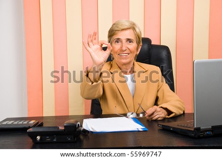 Happy senior business woman showing okay sign hand and sitting on chair at desktop
