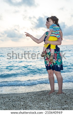 Mother pointing and showing something to her toddler boy on the beach at sunset