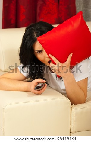 Alone woman laying on sofa watching a horror movie at tv and being scared and covering eye with pillow