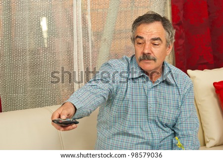 Senior man sitting on couch in living room and opening tv with remote control