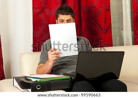 Man working home and sitting on couch using laptop and reading paper
