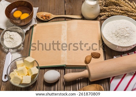 Cooking. Rural vintage wood kitchen table with blank cook book, baking cake ingredients (eggs, flour, milk, butter, sugar) and cooking utensils around. Country background with free recipe text space.