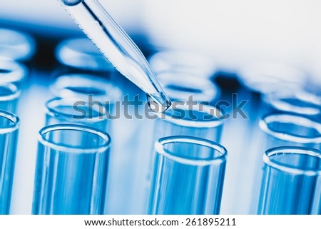 Lab. A close-up of a laboratory glass pipette with emerging drop of substance over one of several test tubes on a light  background