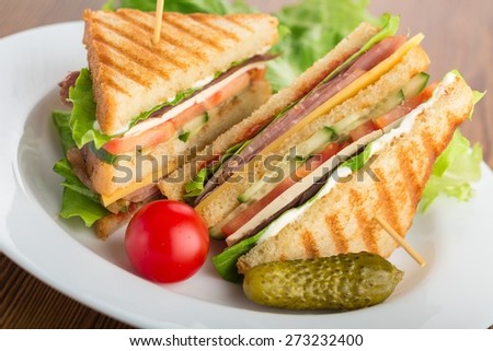 Sandwich. Photo of a club sandwich made with turkey, bacon, ham, tomato, cheese, lettuce, and garnished with a pickle and two cherry tomatoes.