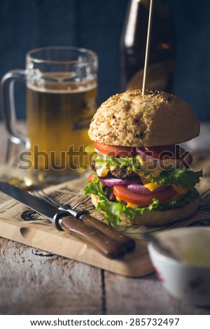 Delicious burger stacked high with a beef patty, cheese, fresh lettuce, onion and tomato on a fresh bun with sesame seed standing on brown paper with glass of beer on a wooden background
