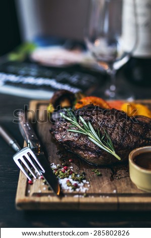 Grilled Steak with salt and pepper on meat cutting board with grilled vegetables and wine on dark wooden background. Bottle of red wine on the background.