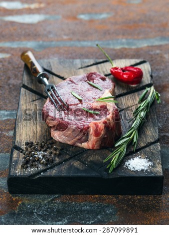 Raw fresh meat ribeye steak, chili pepper, salt and rosemary on cutting board with meat fork on dark background