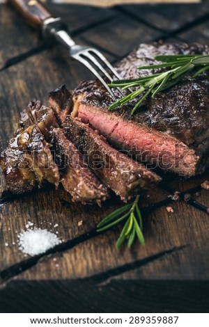 Close up sliced medium rare grilled beef steak with salt, pepper and rosemary on meat cutting board on dark wooden background
