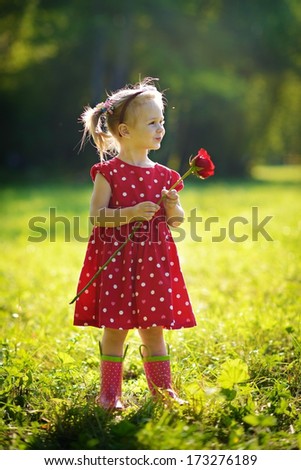 A cute little happy girl holding a red rose in the green meadow on a sunny summer day
