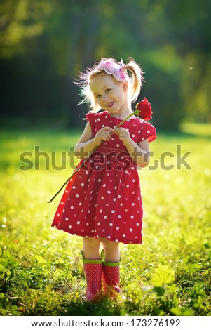 A cute little happy girl holding a red rose in the green meadow on a sunny summer day
