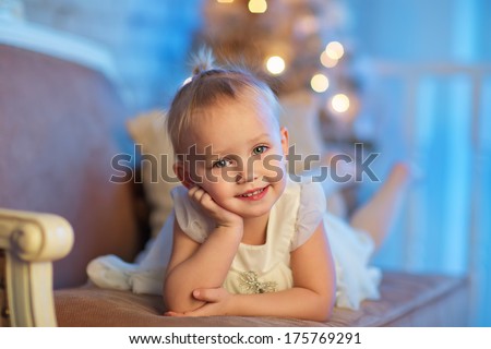 A lovely 3 year old girl in a beautiful white dress lying on a sofa in front of the decorated Christmas tree