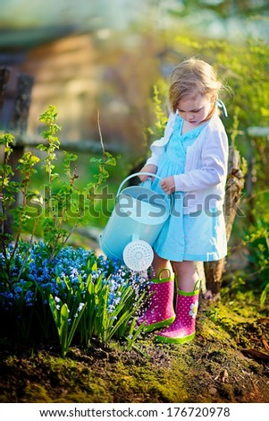 A cute little girl in bright pink rubber boots watering blooming flowers in a garden