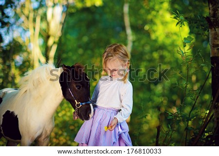 A cute white little girl in jockey boots caressing her little pony in a green forest on a sunny summer day