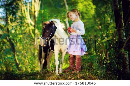 A cute white little girl in jockey boots caressing her little pony in a green forest on a sunny summer day