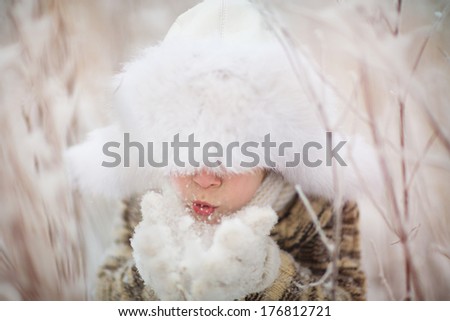 A cute boy in a white fur cap with ear-flaps blowing the snowflakes off his mittens on a frosty winter day