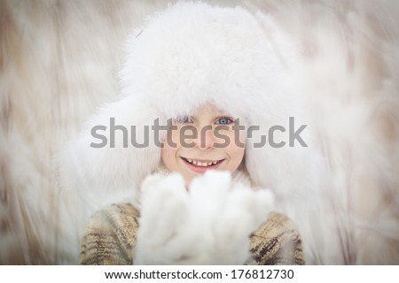 A cute smiling boy in a white fur cap with ear-flaps on a frosty winter day close up