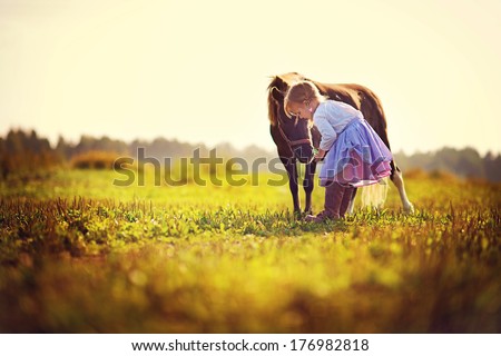 A cute white girl in jockey boots caressing her little pony in the field on a sunny summer day