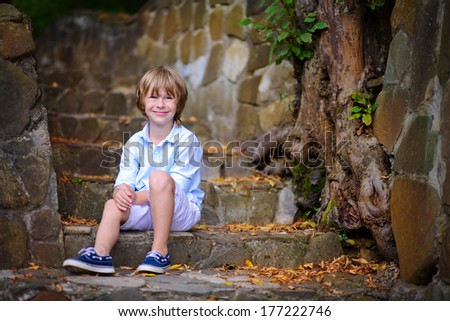 A handsome sad little boy sitting on the stone step against the old stone wall and smiling