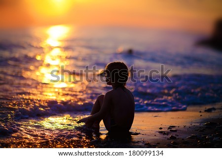 A silhouette of a boy sitting on the sea shore and looking to the sunset