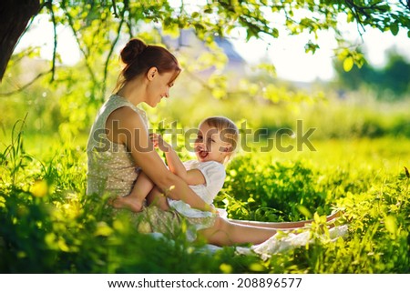 A happy family having a picnic in the green garden in a sunny spring day: a beautiful smiling mother sitting on green grass and her little laughing daughter on her legs