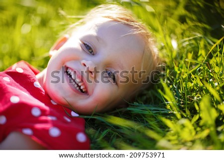 A portrait of a sweet little laughing girl lying on green grass in a sunny summer day