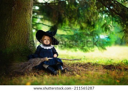 Little cute girl in black witch costume and a magic hat sitting under a tree with a broom in a sunny autumn day. Halloween. National holidays and traditions. Fairy tale. Funny kids.
