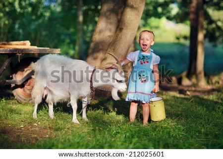A little pretty blond girl in a white dress and blue apron holding a milk-can and playing with her pet goat in the field in a sunny summer day. Kids as helpers in the country.