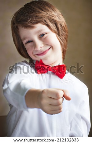 A cheerful cute smiling boy in a white shirt with a red bow-tie holding something you want (flowers, stick lolly) on a grey background closeup. Young handsome gentleman congratulating you with present