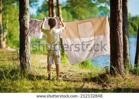 A little cute boy hanging washing on a line in a pine forest on the bank of a lake in a sunny summer day. Kids are playing.