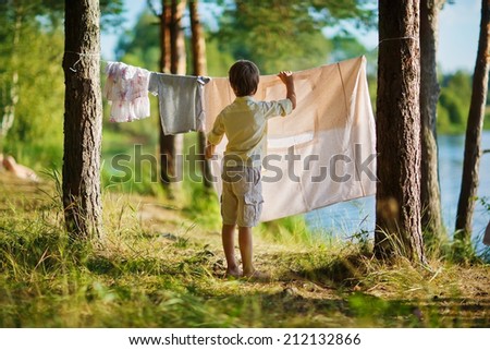 A little cute boy hanging washing on a line in a pine forest on the bank of a lake in a sunny summer day. Kids are playing.