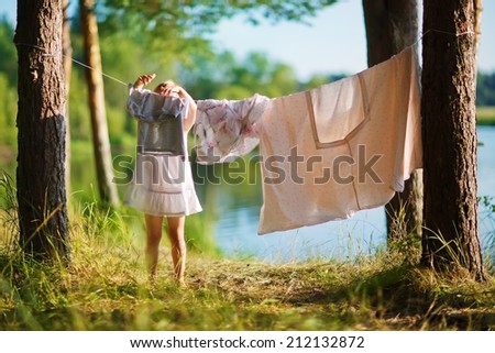 A little cute girl hanging washing on a line in a pine forest on the bank of a lake in a sunny summer day. Kids are playing.