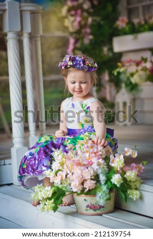 A little sweet girl with a flower diadem near the white beautiful house decorated with flowers in a sunny summer day.