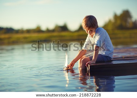 A handsome happy child playing with a tiny boat on water on a sunny summer day. Kids in country. Nature.