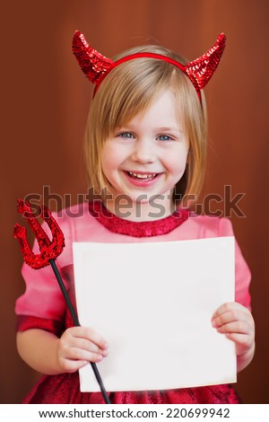 A portrait of a smiling cute little girl disguised as a red devil standing with empty blank in hands, isolated on brown, close up. Holidays concept. Invitation. Funny kids. Masked ball. Halloween.