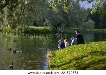 Back view of a father and his little kids sitting on the river shore and looking at ducks on a sunny spring day