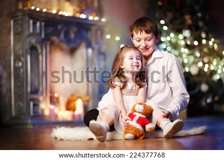 A little happy lovely girl sitting on the floor with her big brother against Christmas Tree and a fireplace. Kids and holidays