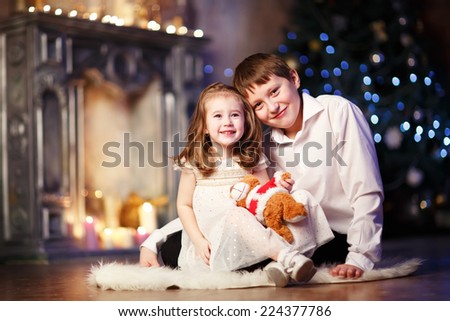 A little happy lovely girl sitting on the floor with her big brother against Christmas Tree and a fireplace and opening Christmas presents wrapped in golden paper with curiosity. Kids and holidays