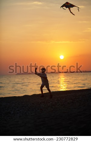 A silhouette of a child running with a kite along the sea coast against the sunset sky on a warm summer evening