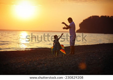 Happy family concept: a young man helps his little daughter to fly a kite on the sea coast against sunset sky and mountains on a warm summer day.