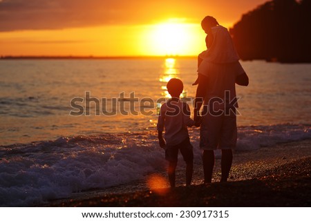 Silhouettes of a happy family: a strong healthy man and his children - little boy and girl standing on the sea shore and looking to the sunset on a warm summer evening
