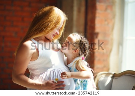 A young pretty pregnant woman with long blonde hair hugging her little cute daughter near the window at home. Happy family concept. Parents and kids relationship.
