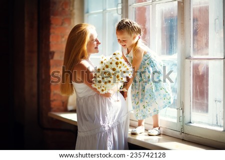 A young pretty pregnant woman with long blonde hair hugging her little cute daughter standing on the windowsill at home. Happy family concept. Parents and kids relationship.