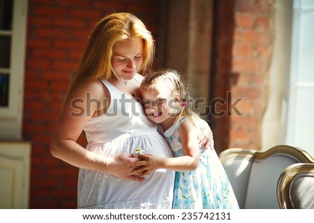 A young pretty pregnant woman with long blonde hair hugging her little cute daughter near the window at home. Happy family concept. Parents and kids relationship.
