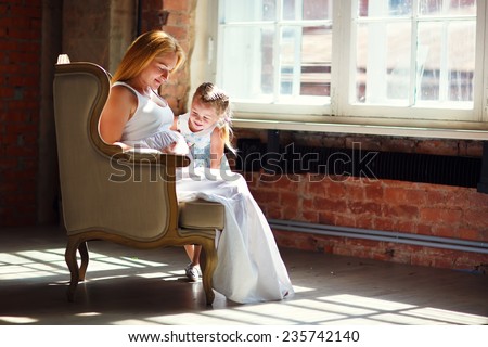 A young pretty pregnant woman with long blonde hair sitting in a chair opposite the window and hugging her little cute daughter. Happy family concept. Parents and kids relationship.